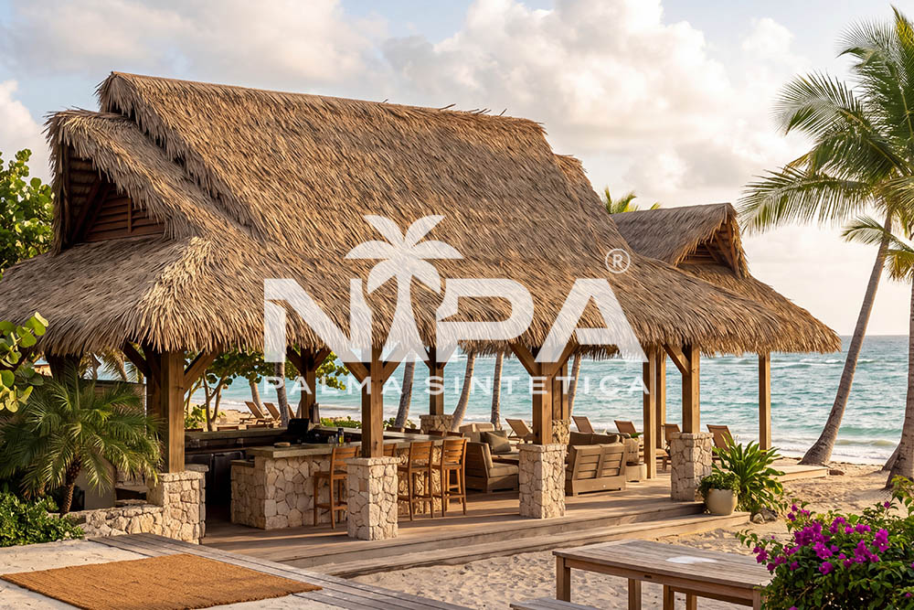 Restaurante de playa con cubierta de palma sintética de varias aguas, columnas de madera sobre bases de piedra y el mar al fondo