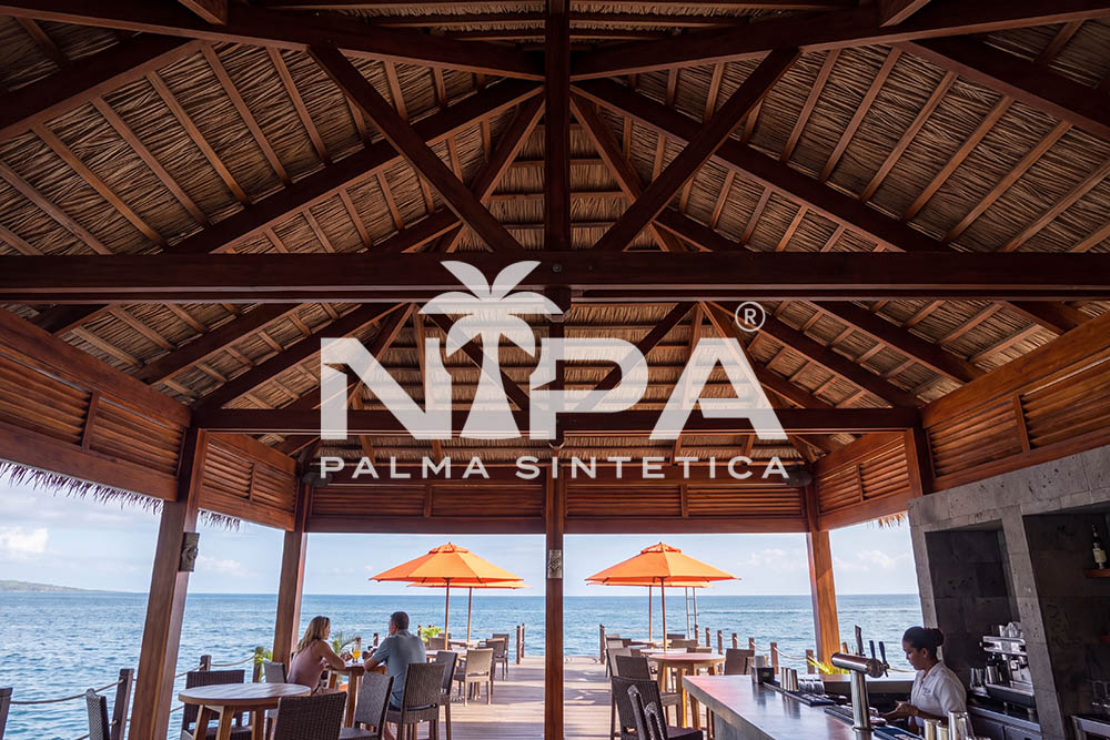 Restaurant with a bar under a synthetic palm thatch roof, exposed timber trusses and the sea in the background