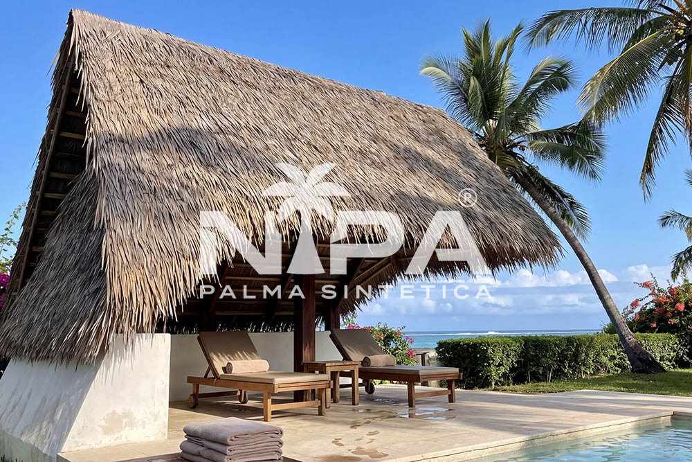 Gable synthetic palm thatch roof coming down to a white wall, over sun loungers by a pool
