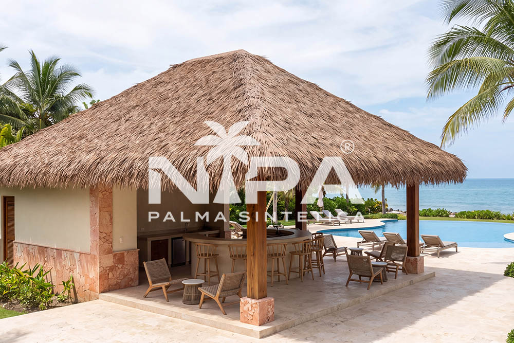 Bar de piscina con cubierta de palma sintética a cuatro aguas, columnas de piedra rosada, barra curva y el mar al fondo