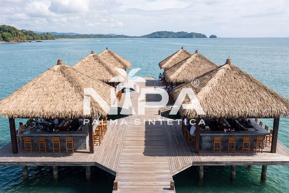 Vista aérea de seis kioscos cónicos de palma sintética con barras, sobre un muelle de madera en el mar