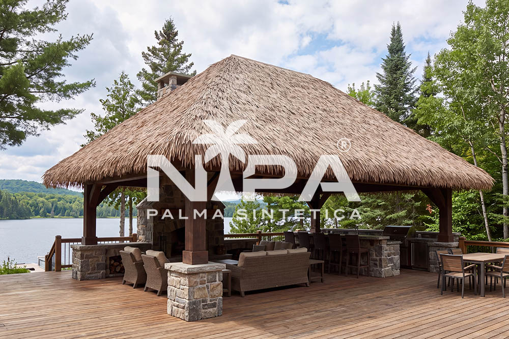 Pabellón de palma sintética con chimenea de piedra que atraviesa la cubierta, sala y barra sobre una terraza de madera junto a un lago