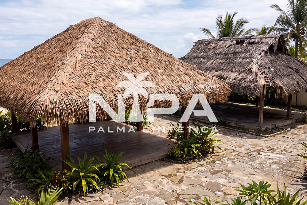 Hipped synthetic palm thatch palapa on a timber platform; in the background, another grayer, weathered roof